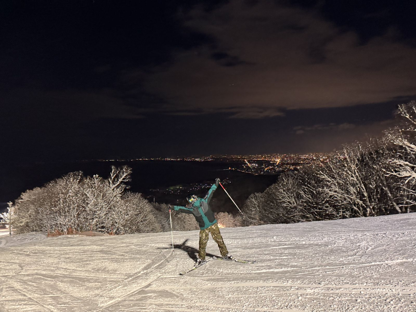 夜滑時在 Onze 雪道上張開雙手的滑雪者,背後是海岸線燈火