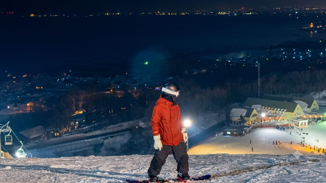 夜晚在 Onze 山頂準備滑落的單板學員，背後是石狩灣沿岸燈火和雪場大樓